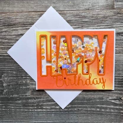 Colorful handmade birthday card with sequins and orange lettering on wooden background.