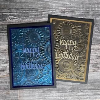 Two handmade birthday cards with gear patterns, one in blue and one in gold, on a wooden surface.